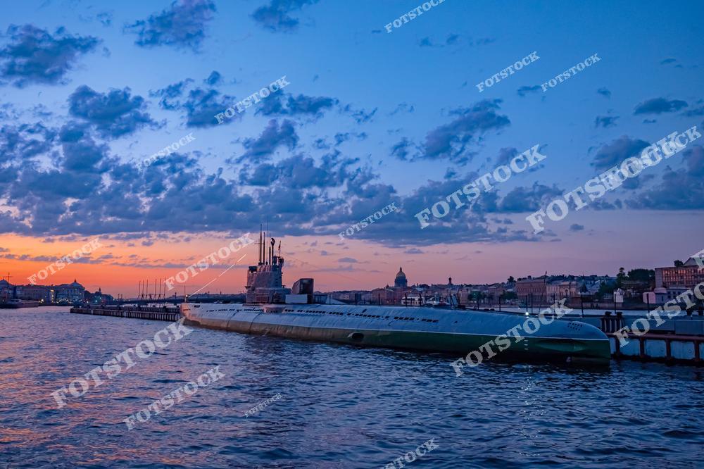 На фотографии изображена подводная лодка "Народоволец", являющаяся экспонатом Музея подводных лодок Санкт-Петербурга. Этот исторический памятник расположен на Неве и входит в состав Центрального военно-морского музея. Лодка отражает значимые этапы развития подводного флота России. Закатное небо и городские силуэты подчеркивают уникальность расположения музея.